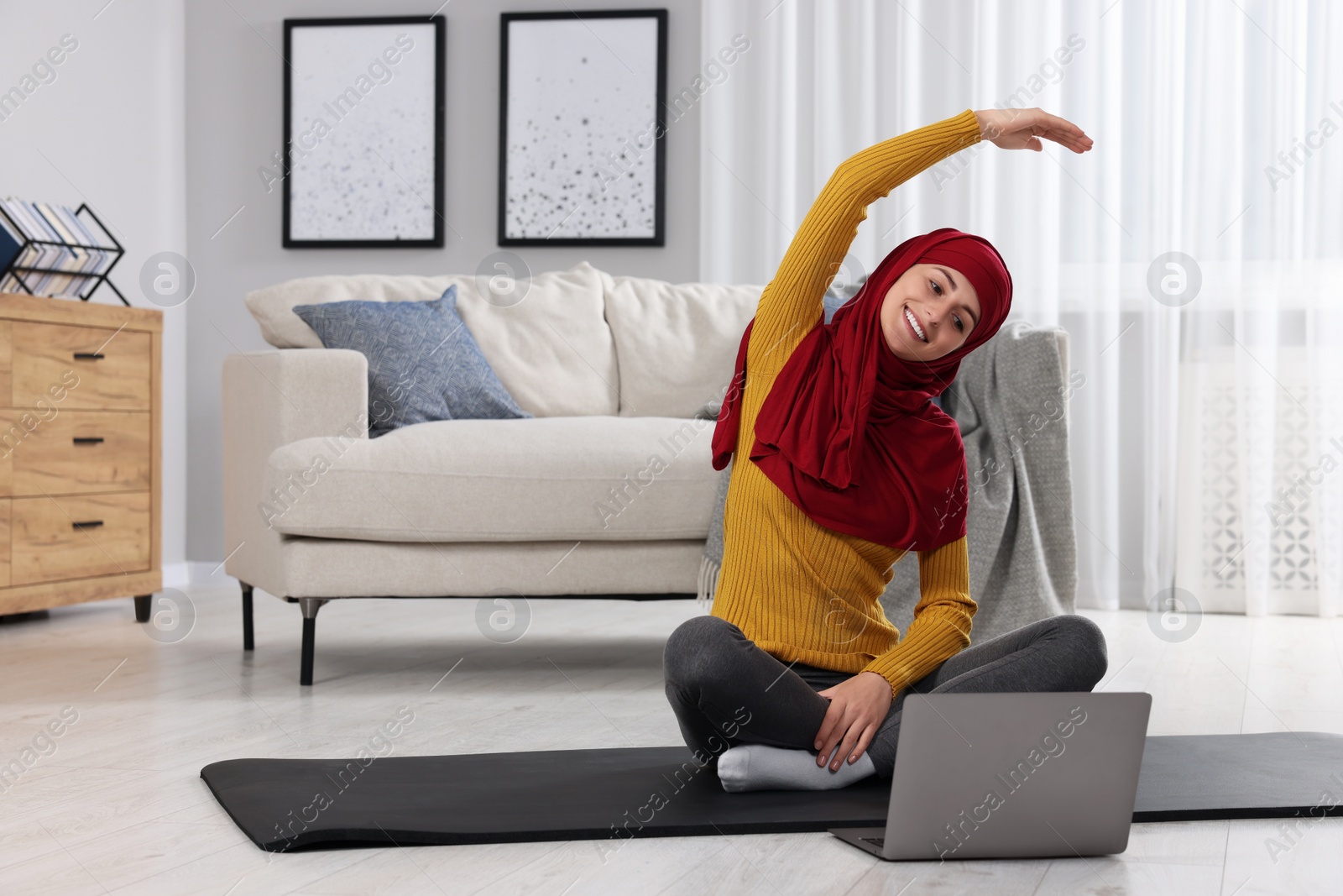 Photo of Muslim woman in hijab doing exercise near laptop on fitness mat at home. Space for text