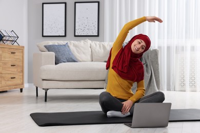 Muslim woman in hijab doing exercise near laptop on fitness mat at home. Space for text