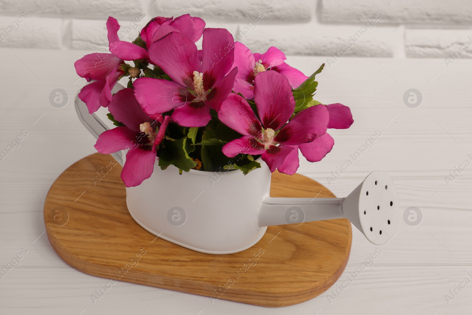 Photo of Beautiful flowers in watering can on white wooden table near brick wall