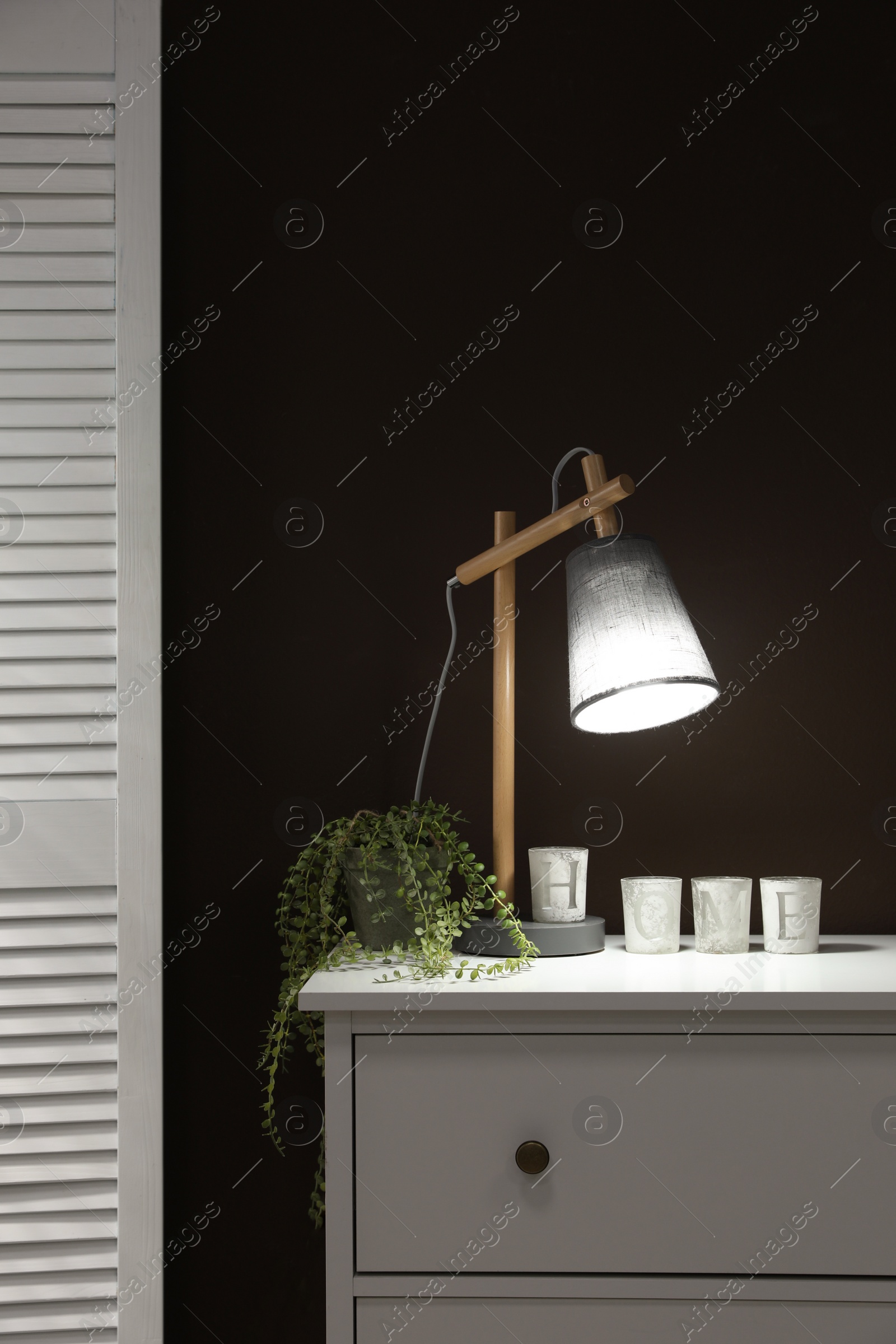 Photo of Stylish lamp, decor and green plant on white chest of drawers near brown wall indoors. Interior design