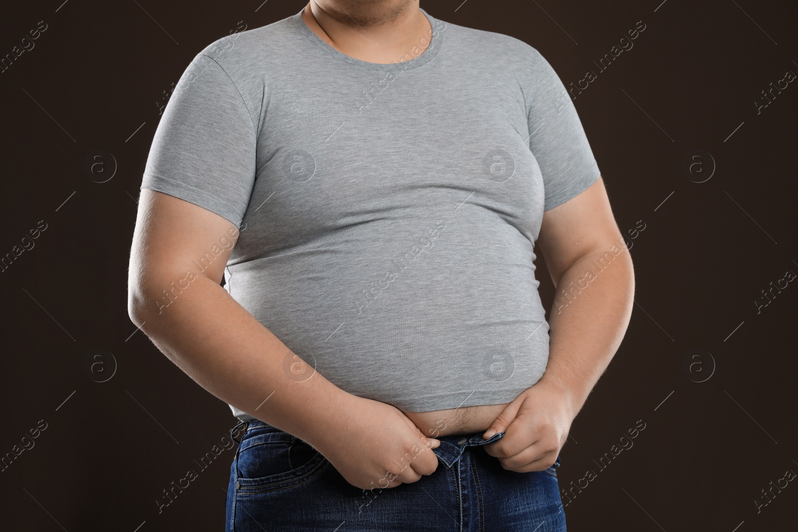 Photo of Overweight man trying to button up tight jeans on dark brown background, closeup