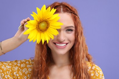 Photo of Beautiful young hippie woman covering eye with sunflower on violet background