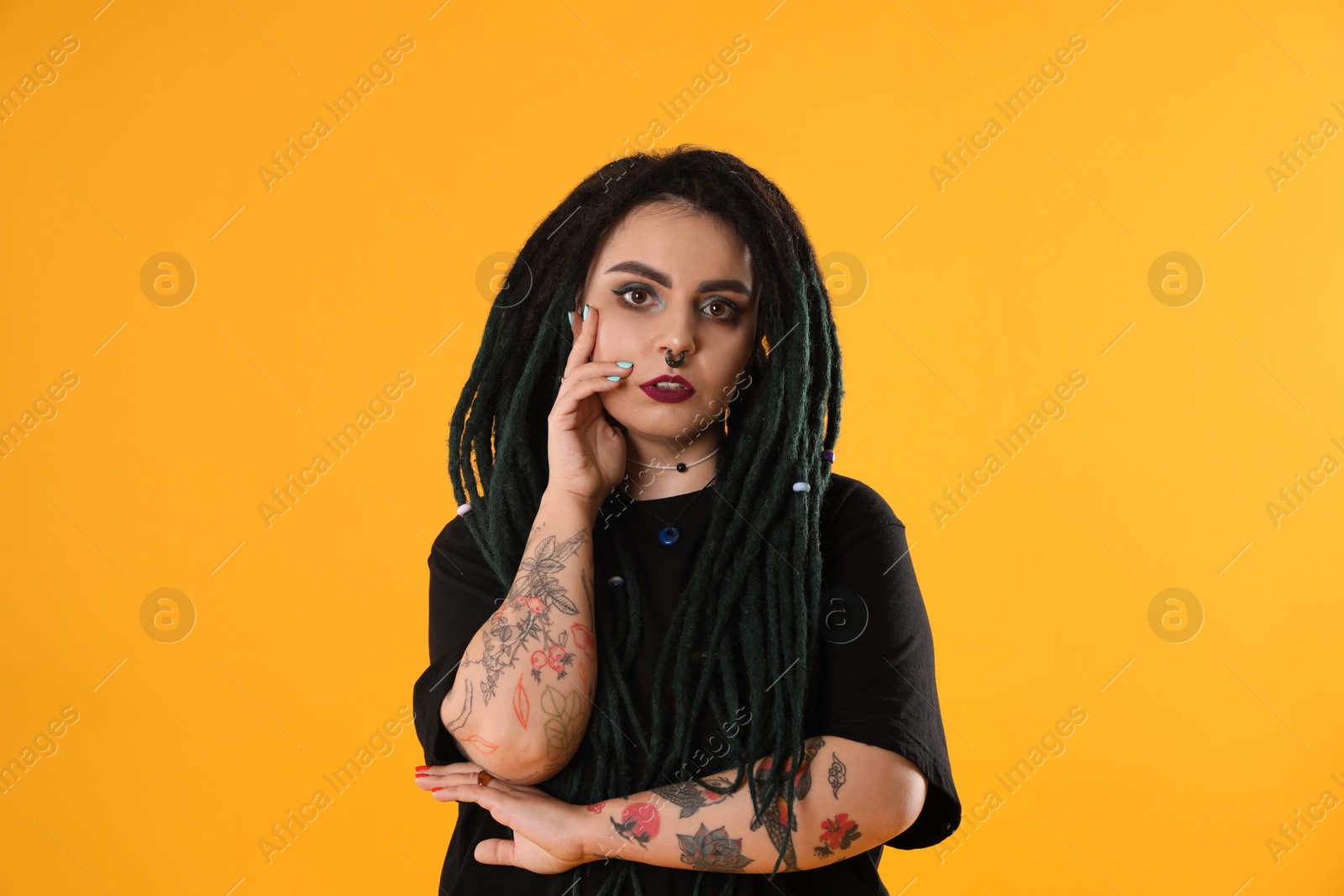 Photo of Beautiful young woman with tattoos on arms, nose piercing and dreadlocks against yellow background