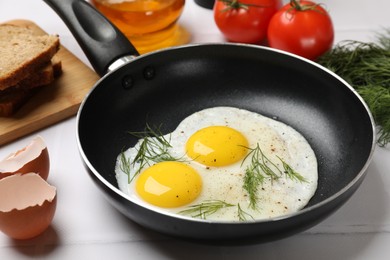 Frying pan with tasty cooked eggs, dill and other products on white tiled table, closeup