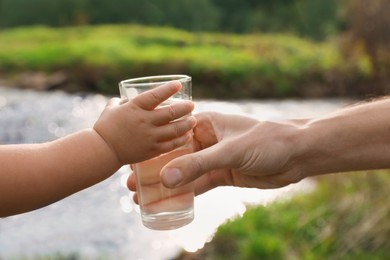 Father giving her daughter glass of fresh water near stream on sunny day, closeup