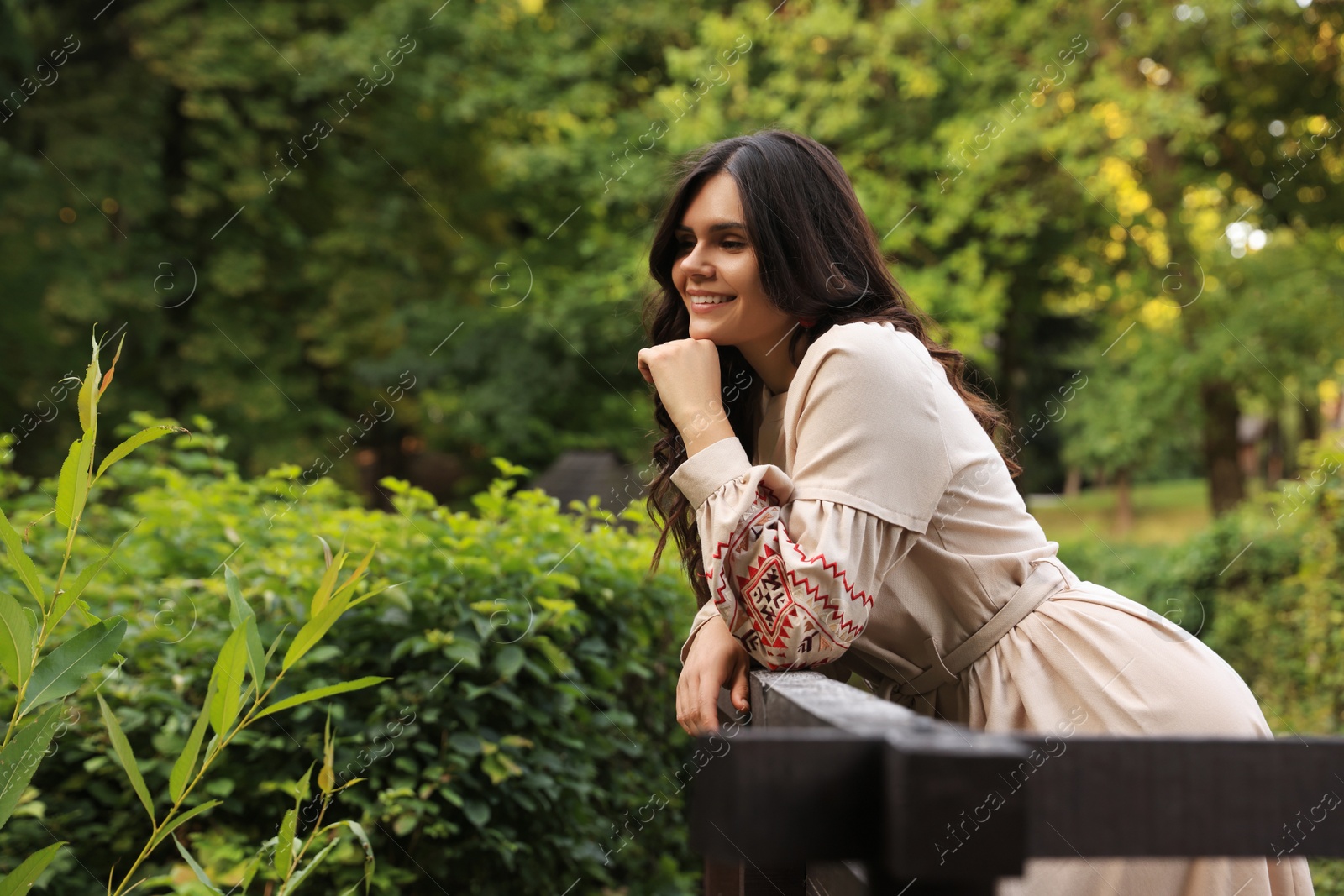 Photo of Beautiful woman wearing embroidered dress near wooden railing in countryside, space for text. Ukrainian national clothes