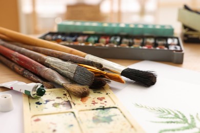 Photo of Different brushes, colorful paints, palette and drawing on table indoors, closeup. Artist's workplace