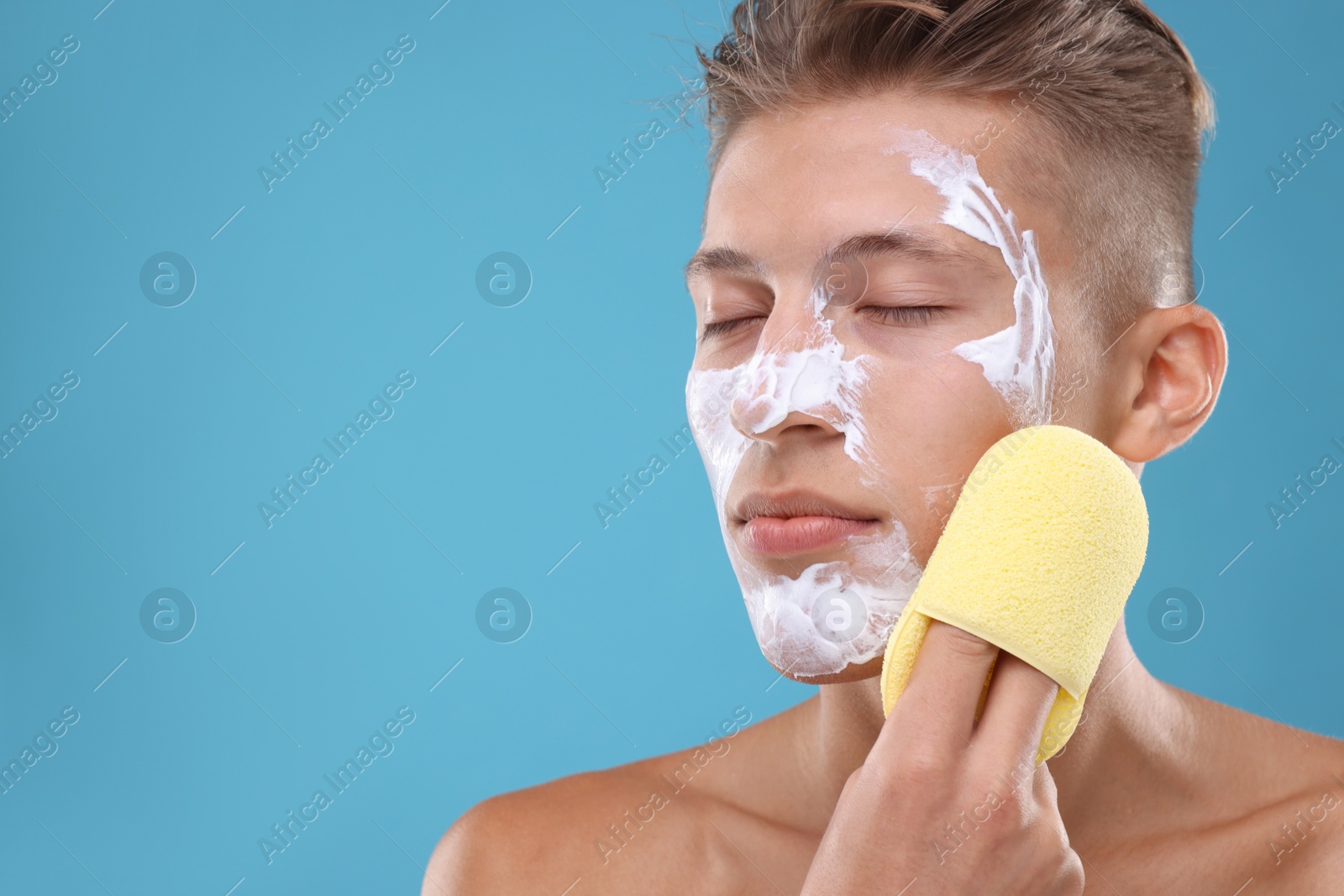 Photo of Young man washing off face mask with sponge on light blue background. Space for text
