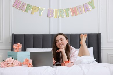 Photo of Beautiful young woman with laptop near gift boxes and rose flowers on bed in room. Happy Birthday
