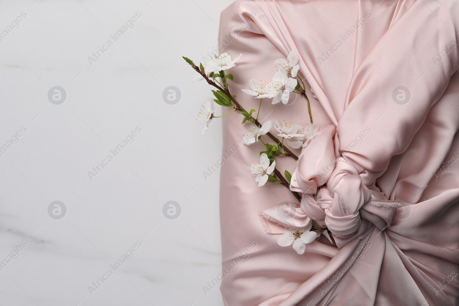 Photo of Furoshiki technique. Gift packed in pink fabric and beautiful flowers on white marble table, top view. Space for text