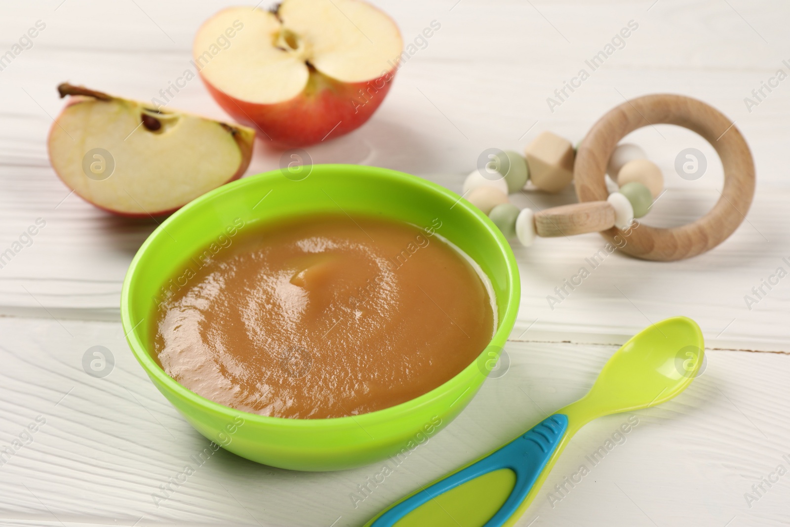 Photo of Baby food. Puree of apples in bowl, spoon and toy on white wooden table