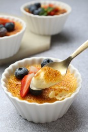 Taking delicious creme brulee with berries from bowl at grey textured table, closeup