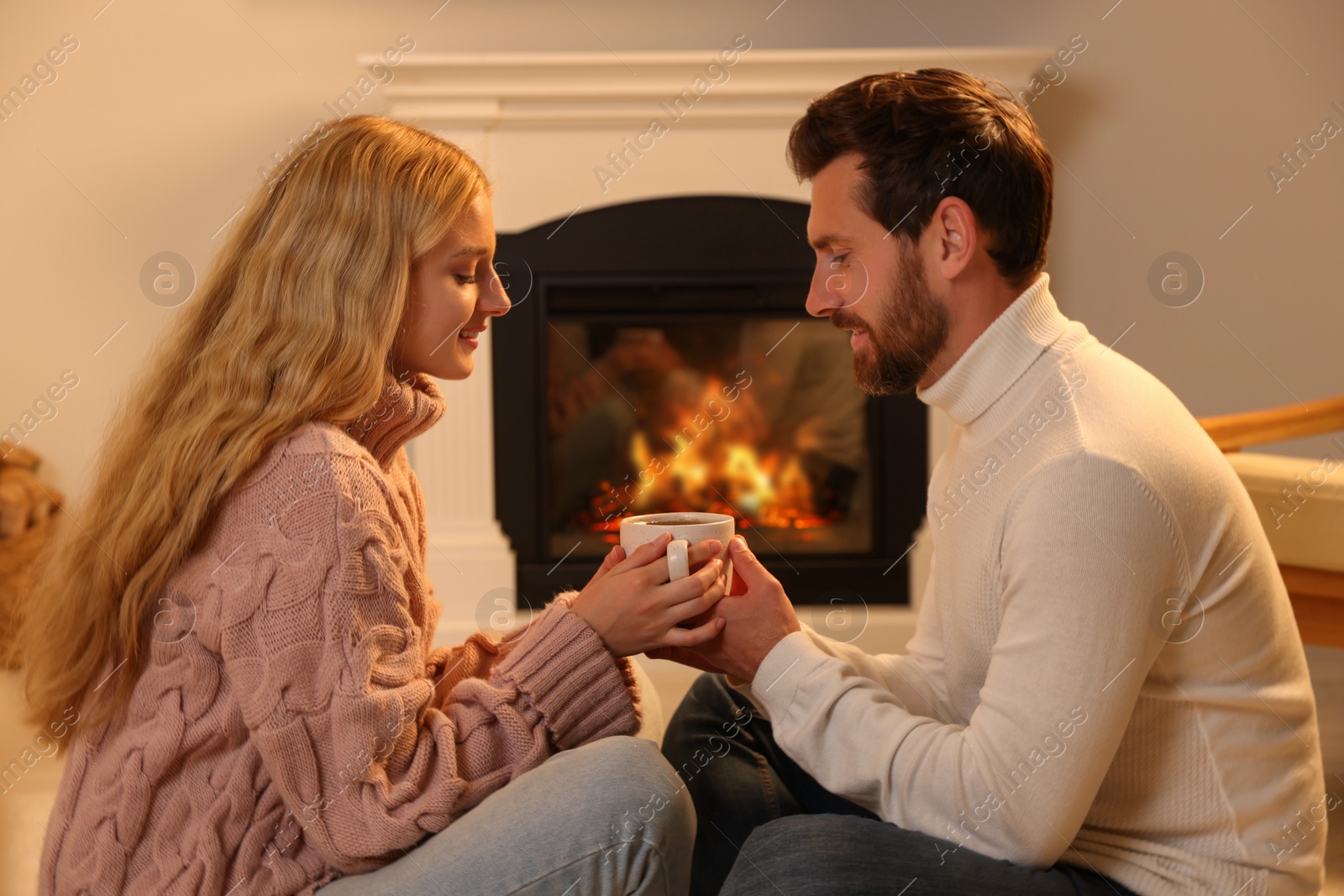 Photo of Lovely couple with cup of hot drink spending time together near fireplace at home