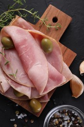 Photo of Tasty ham with olives, garlic, thyme and spices on black textured table, flat lay