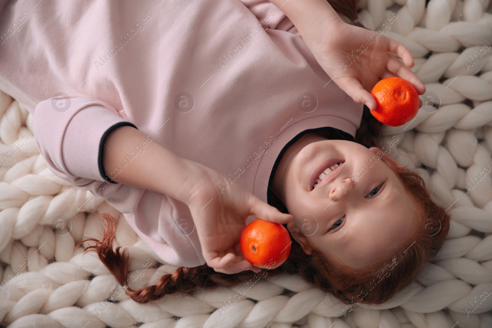 Photo of Happy girl with fresh tangerines lying on knitted plaid, closeup view