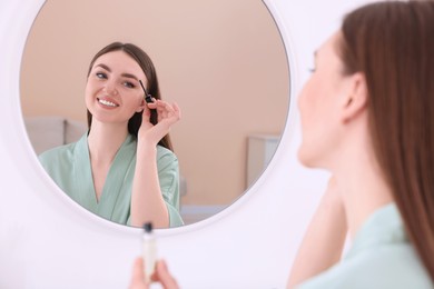Photo of Beautiful young woman applying eyebrow gel near mirror indoors