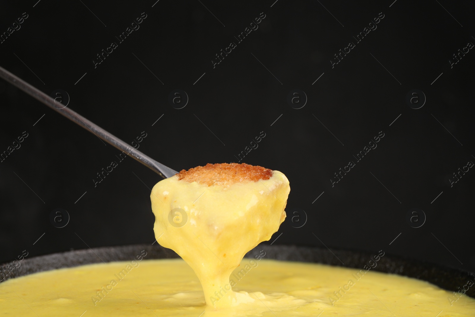 Photo of Dipping piece of bread into fondue pot with tasty melted cheese against dark gray background, closeup