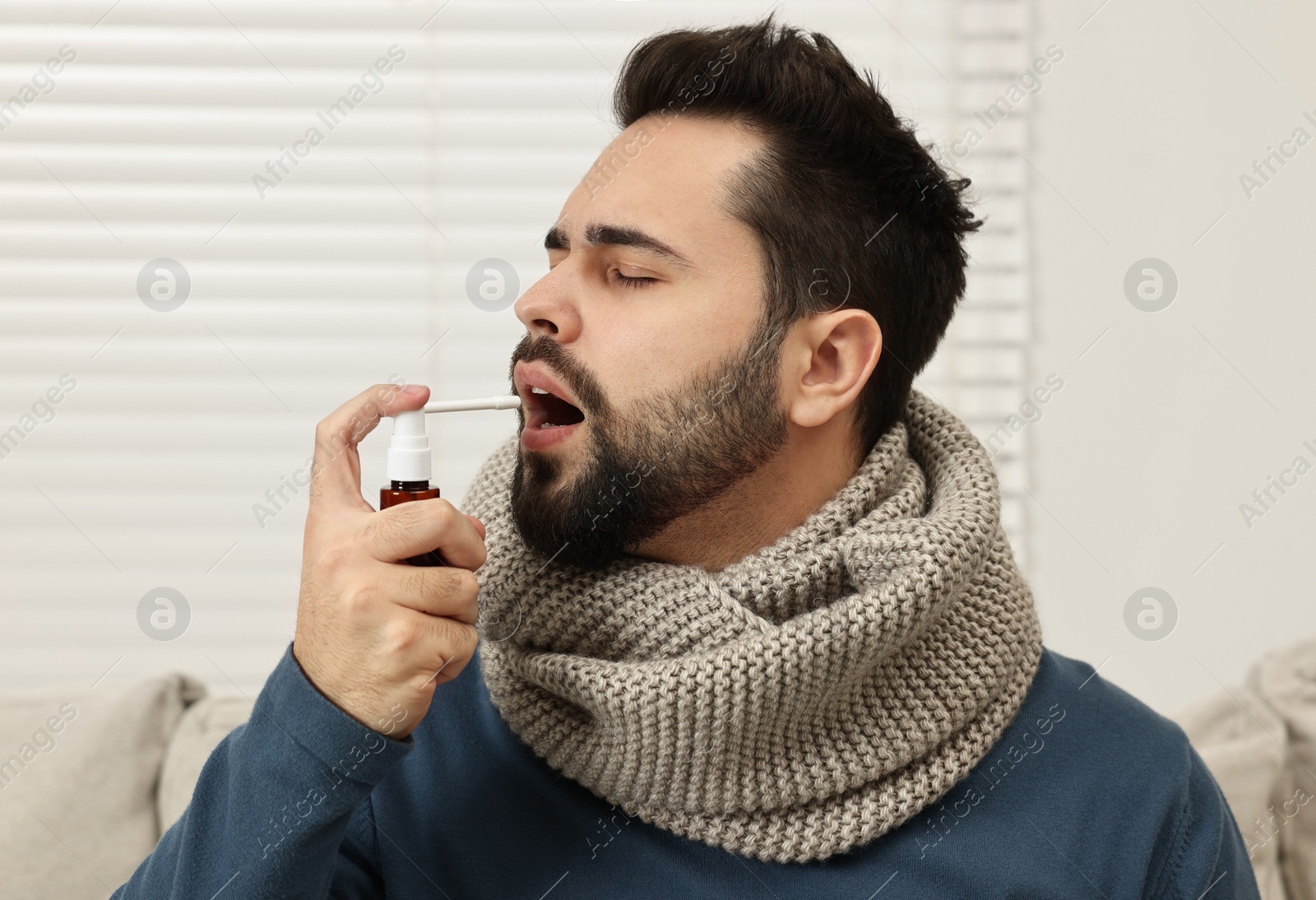 Photo of Young man with scarf using throat spray indoors