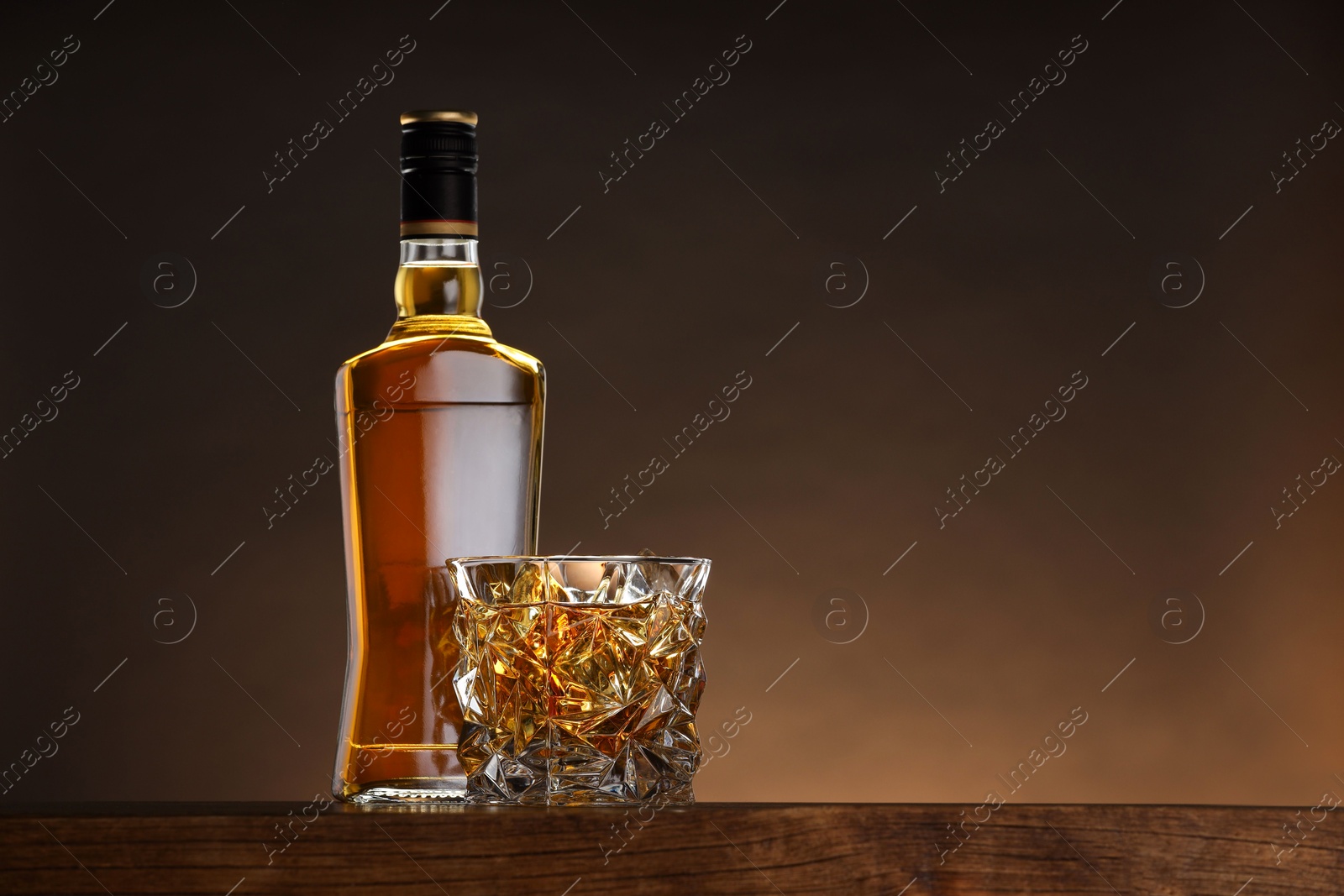 Photo of Whiskey in glass and bottle on wooden table against brown background, space for text