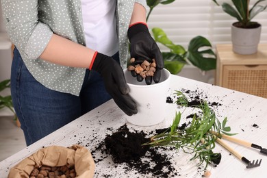 Woman in gloves filling flowerpot with drainage at white table indoors, closeup. Repotting houseplants