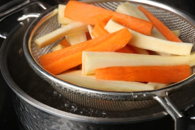 Sieve with cut parsnips and carrots over pot of boiling water, closeup