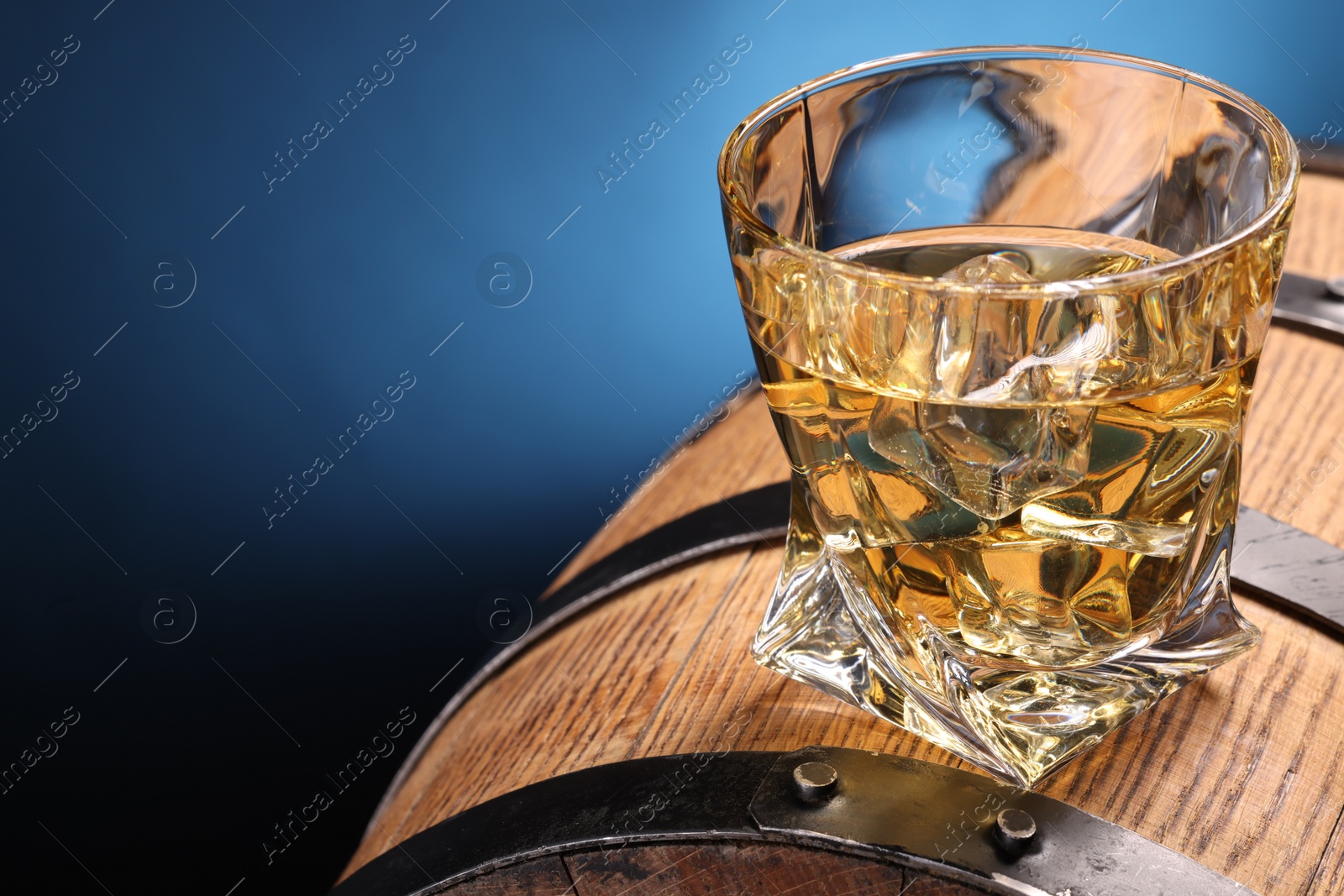 Photo of Whiskey with ice cube in glass on wooden barrel against blue background, closeup. Space for text