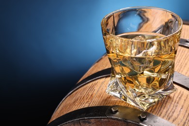 Whiskey with ice cube in glass on wooden barrel against blue background, closeup. Space for text