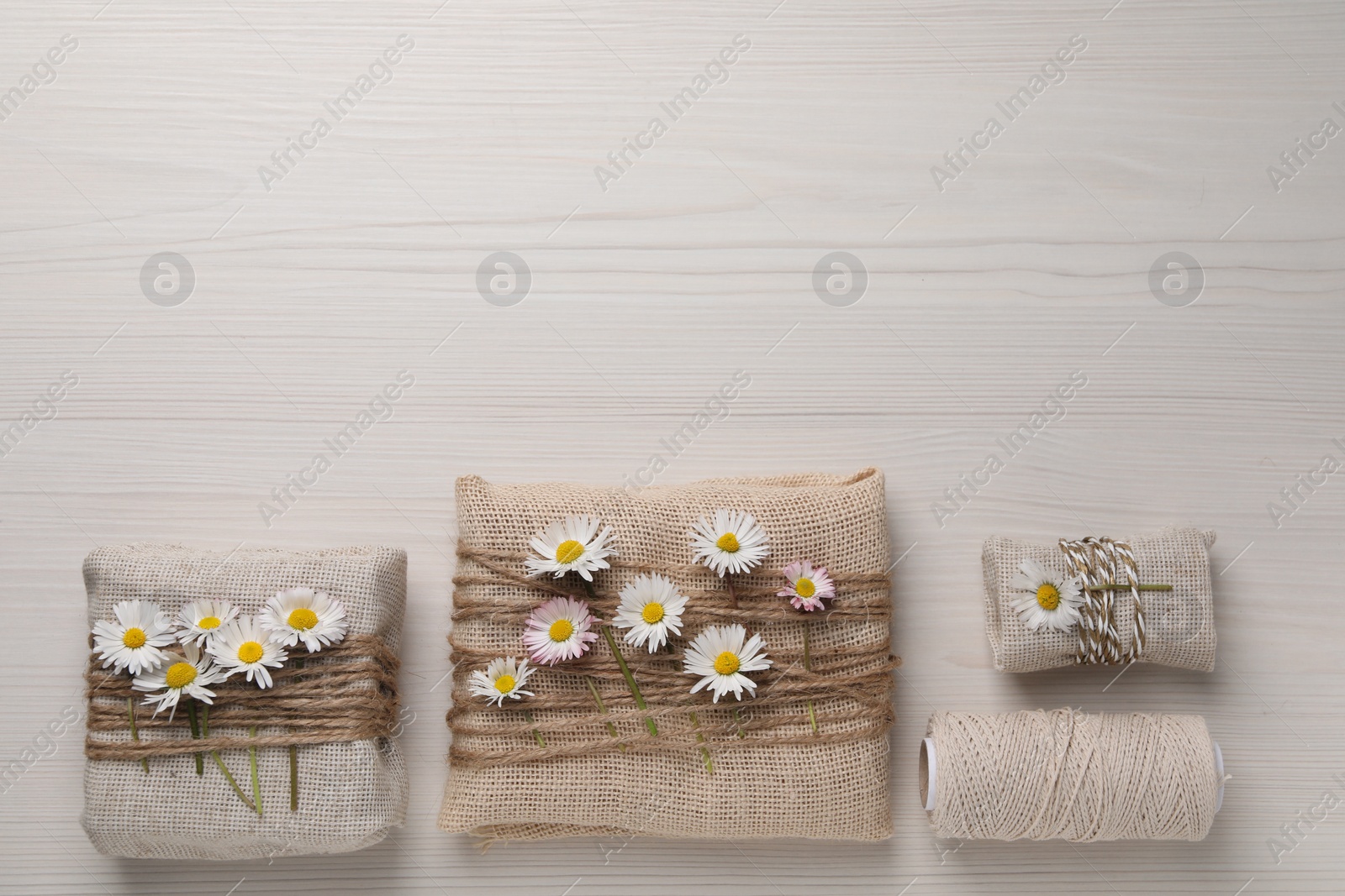 Photo of Gifts packed in burlap fabric with beautiful chamomiles, ropes and spool of threads on white wooden table, flat lay. Space for text