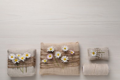 Gifts packed in burlap fabric with beautiful chamomiles, ropes and spool of threads on white wooden table, flat lay. Space for text