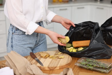 Photo of Garbage sorting. Woman putting food waste into plastic bag at wooden table indoors, closeup