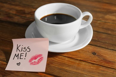 Photo of Sticky note with phrase Kiss Me and cup of coffee on wooden table
