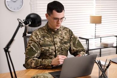Photo of Military service. Young soldier working at wooden table in office