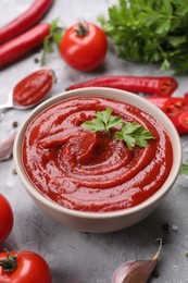 Photo of Organic ketchup in bowl and ingredients on grey textured table, closeup. Tomato sauce