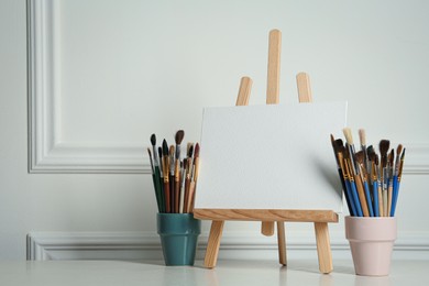 Easel with blank canvas and brushes on white wooden table