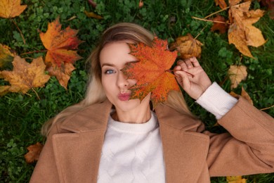 Beautiful woman lying on grass and covering eye with autumn leaf, top view