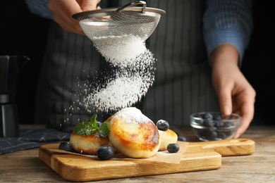 Photo of Woman sieving sugar powder on cottage cheese pancakes at wooden table, closeup