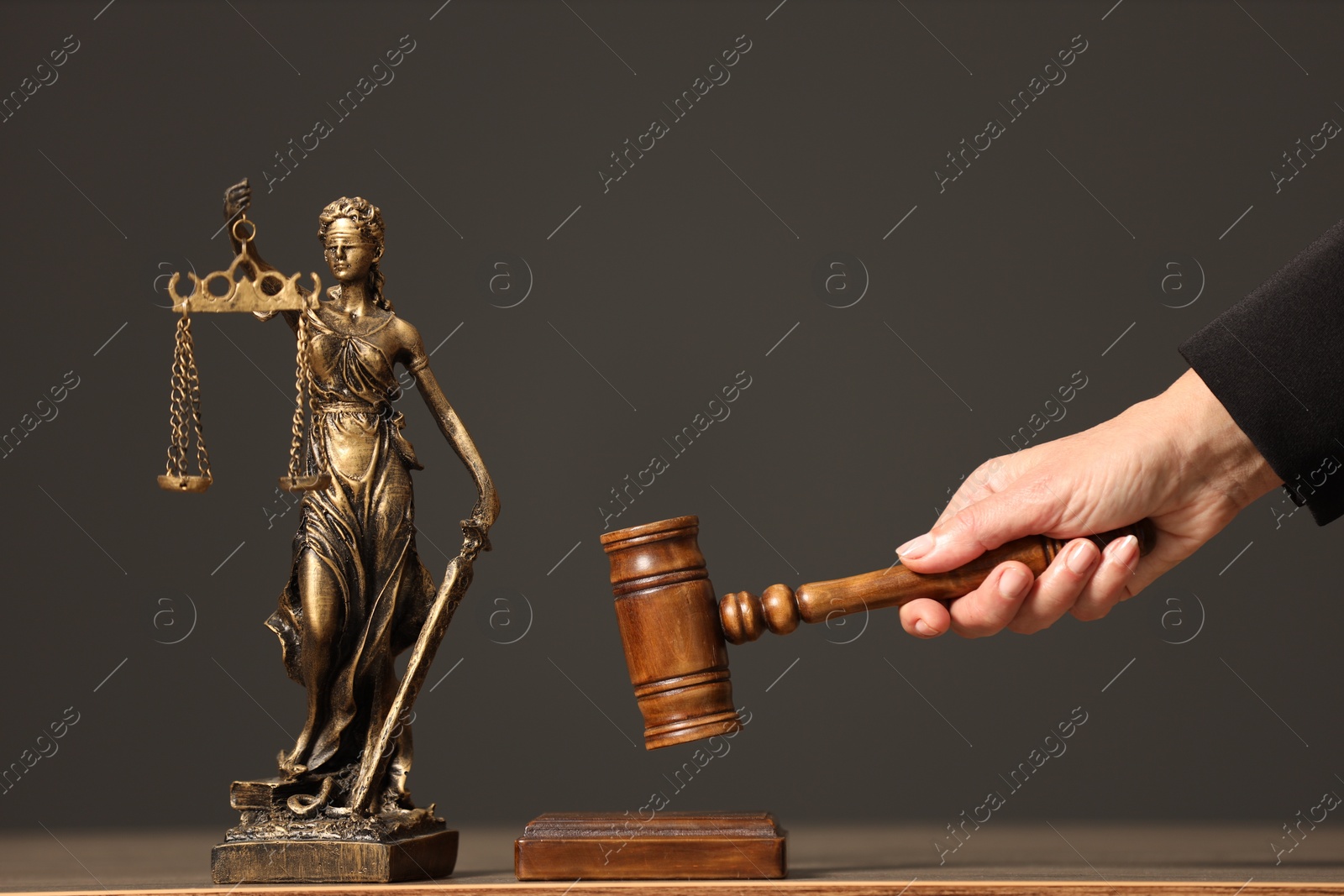 Photo of Judge striking mallet at wooden table against grey background, closeup. Figure of Lady Justice indoors