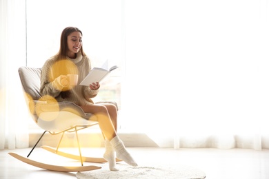 Young woman with cup of coffee reading book near window at home, space for text