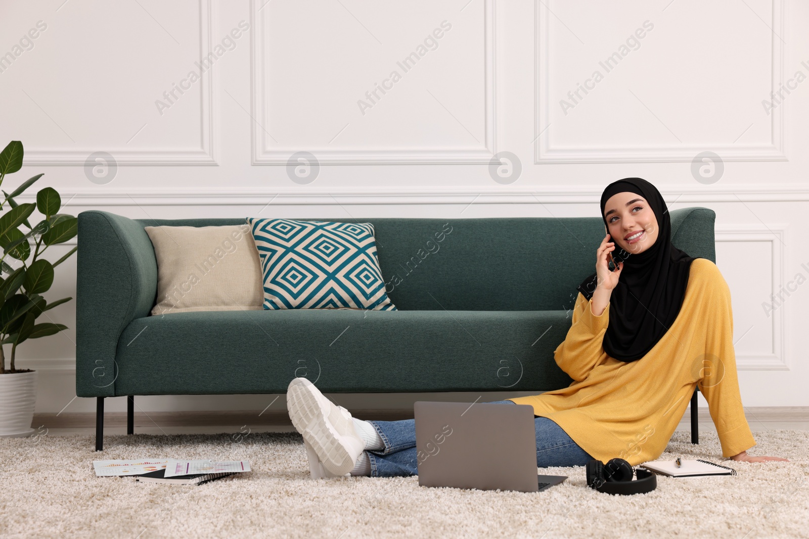 Photo of Muslim woman in hijab talking on smartphone near laptop on floor in room. Space for text