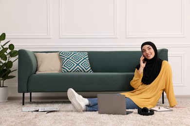 Muslim woman in hijab talking on smartphone near laptop on floor in room. Space for text