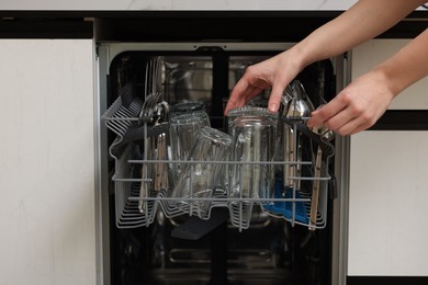 Woman loading dishwasher with glass and cutlery indoors, closeup