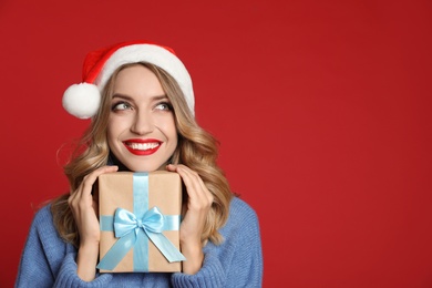 Happy young woman wearing Santa hat with Christmas gift on red background, space for text
