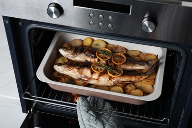Photo of Woman taking out baking tray with sea bass fish and potatoes from oven