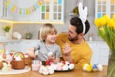 Photo of Father and his cute son having fun while painting Easter eggs at table in kitchen