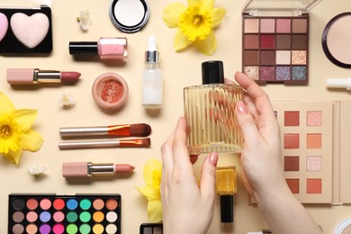 Woman with bottle of perfume, other different makeup products and beautiful spring flowers on beige background, top view