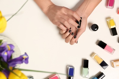 Woman applying nail polish near bottles on white table with flowers, top view