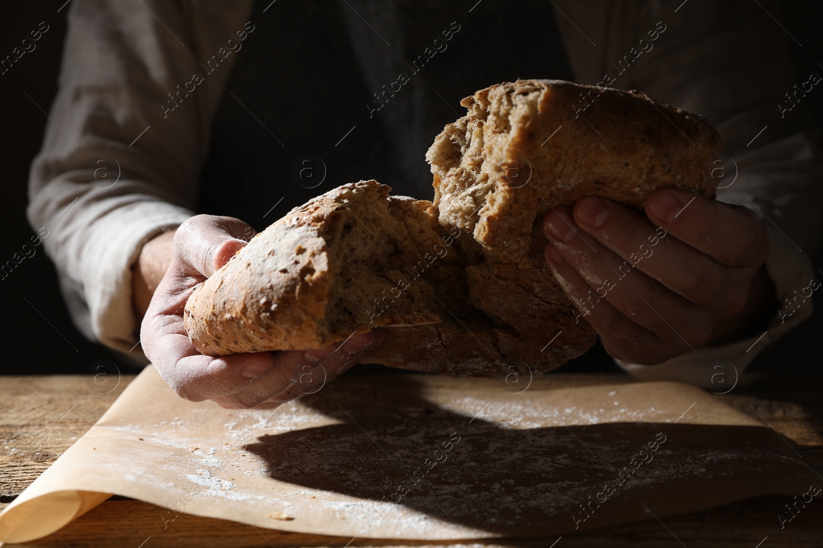 Photo of Man breaking loaf of fresh bread at wooden table, closeup