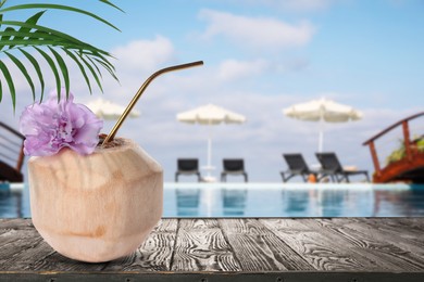 Image of Tasty cocktail in coconut with straw and beautiful flower on wooden table near outdoor swimming pool, space for text