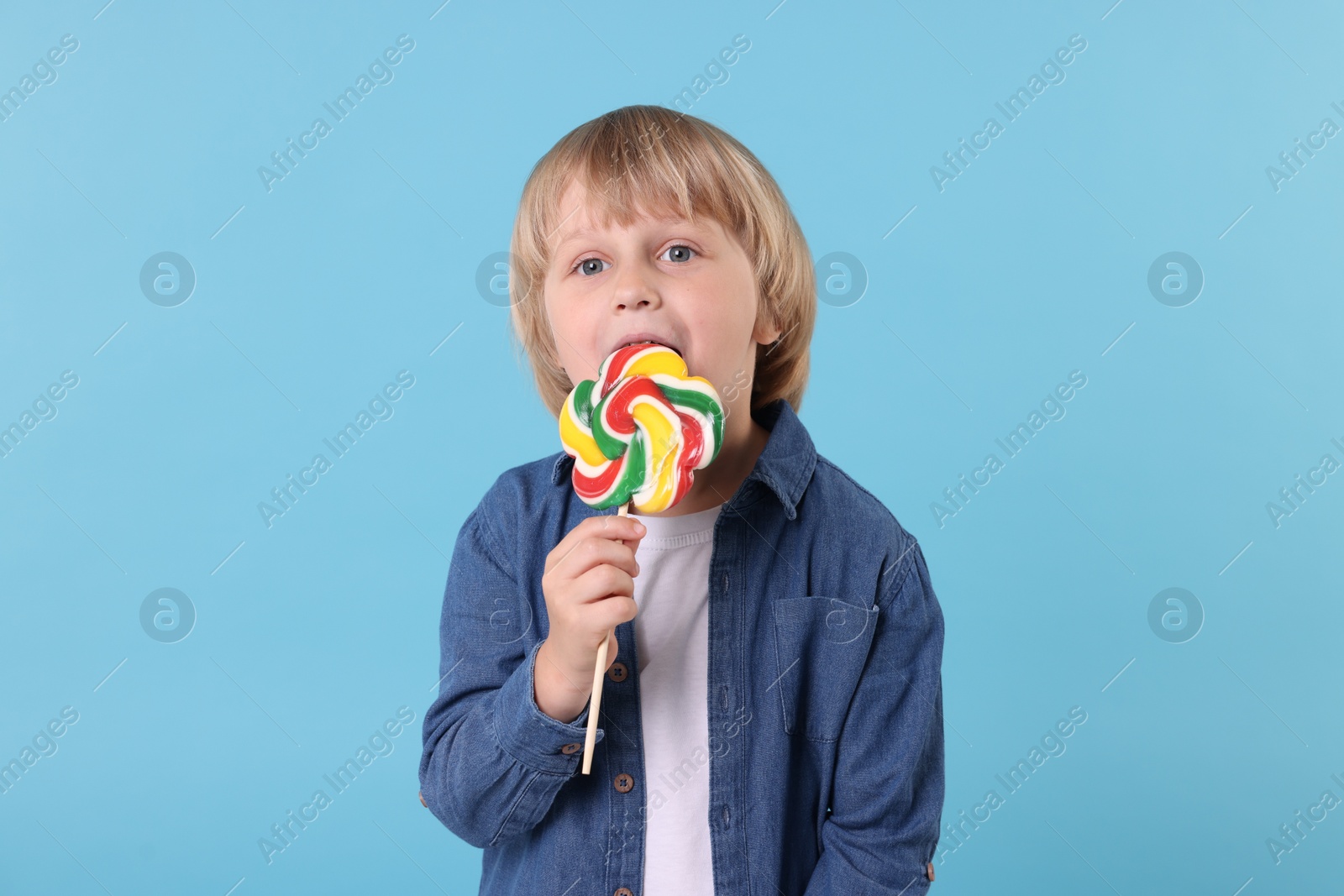 Photo of Cute little boy licking colorful lollipop swirl on light blue background