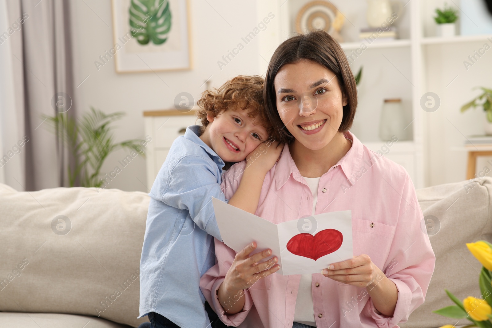 Photo of Little son congratulating his mom with Mother`s day at home. Woman holding handmade greeting card